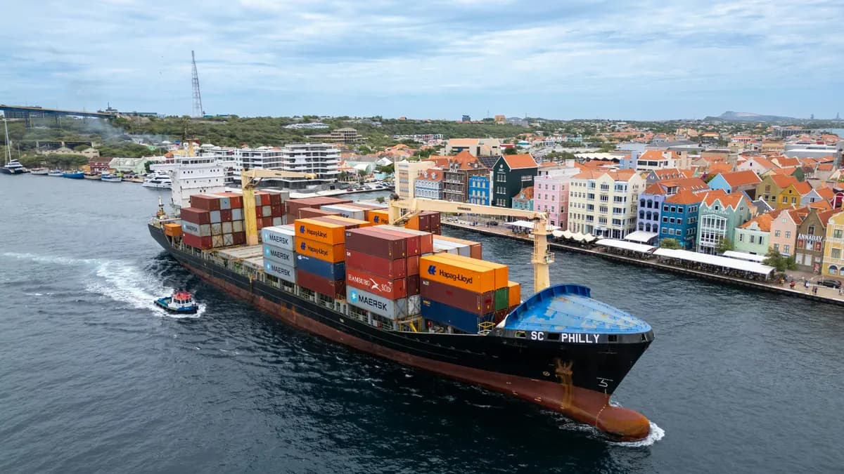 A container ship navigates through the port of Willemstad, Curaçao, with colorful Dutch colonial buildings lining the waterfront.